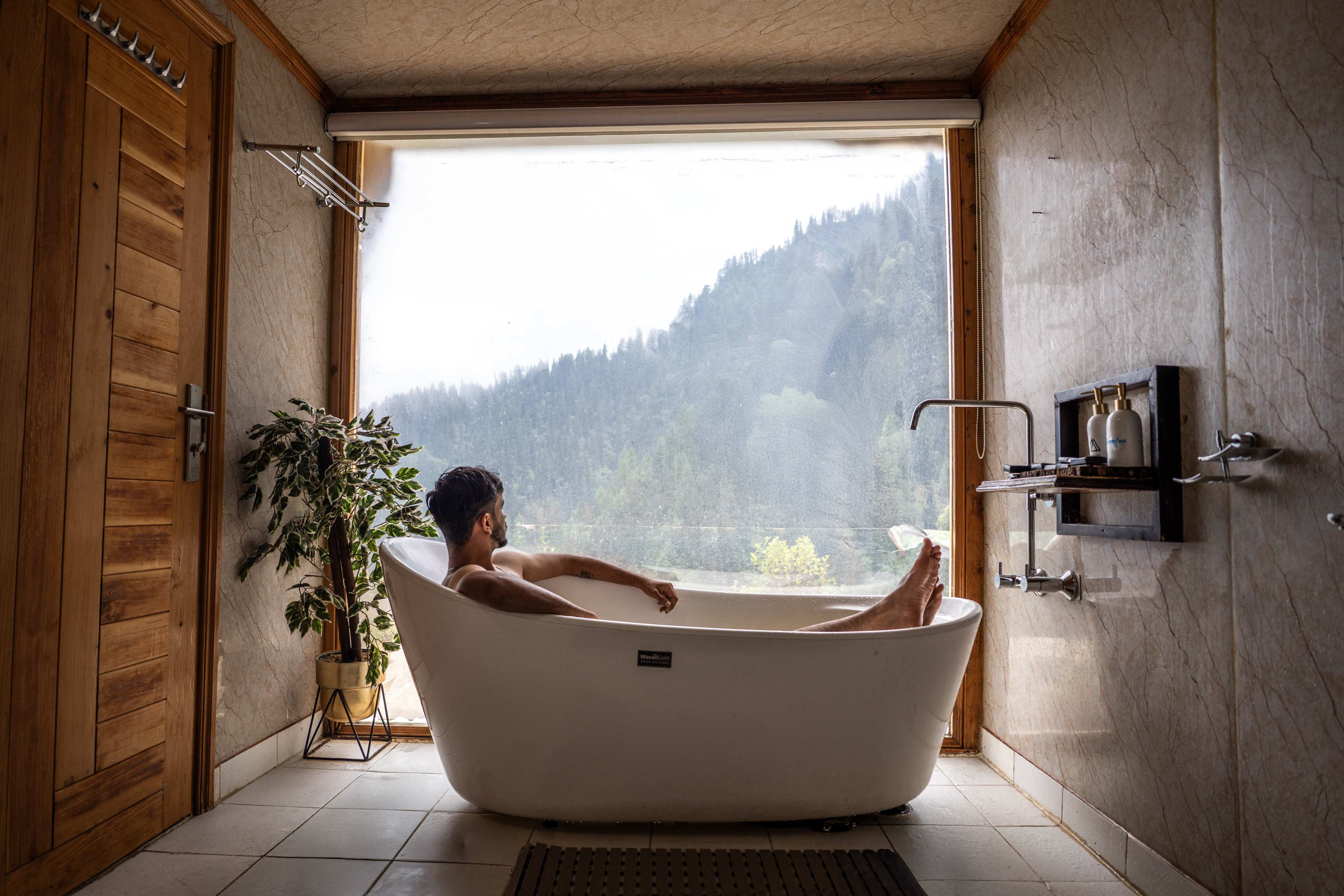 A guest soaking in a freestanding bathtub before a wall of glass, misty forest beyond