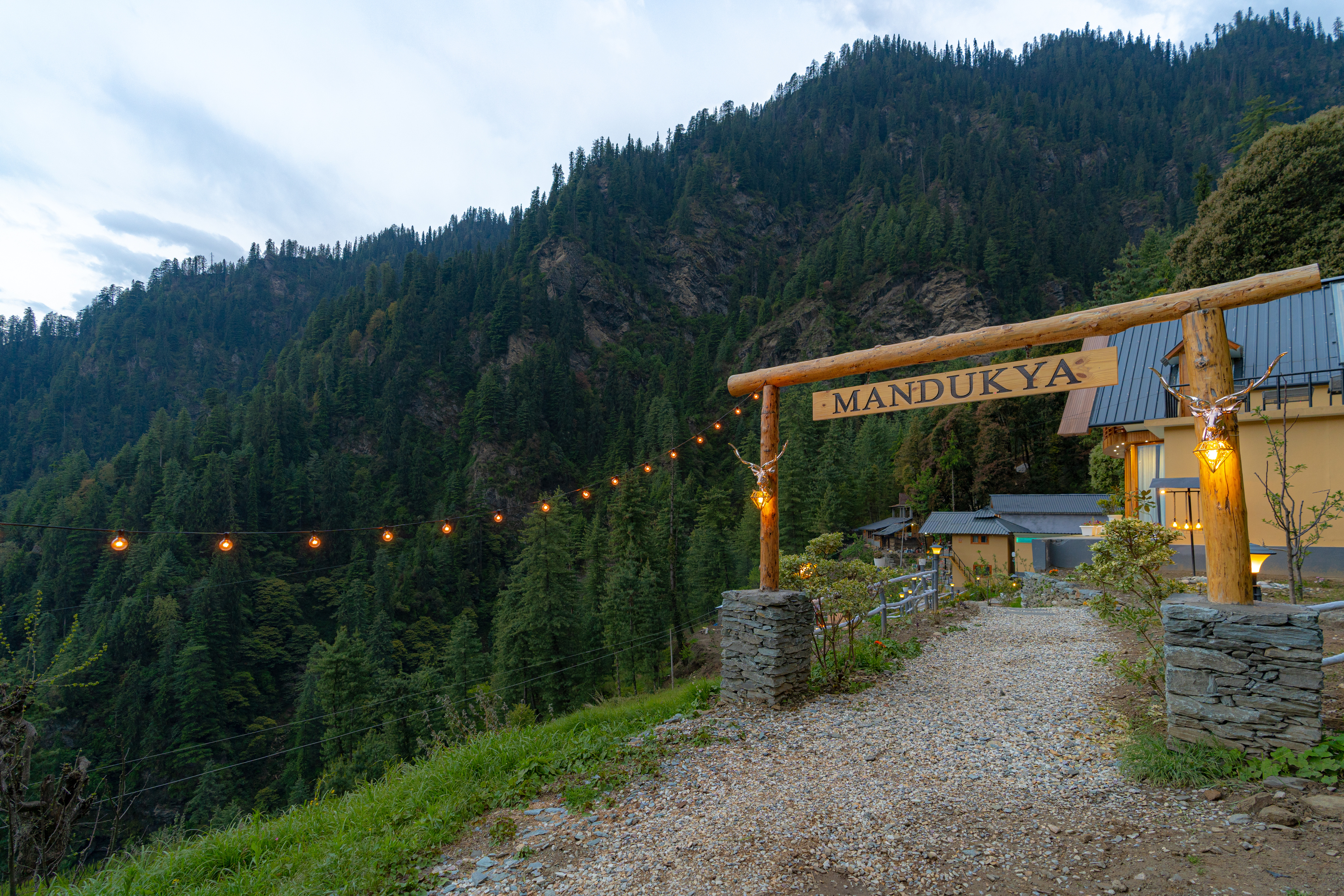 The wooden Mandukya gate at dusk, with a string of lights tracing the hillside