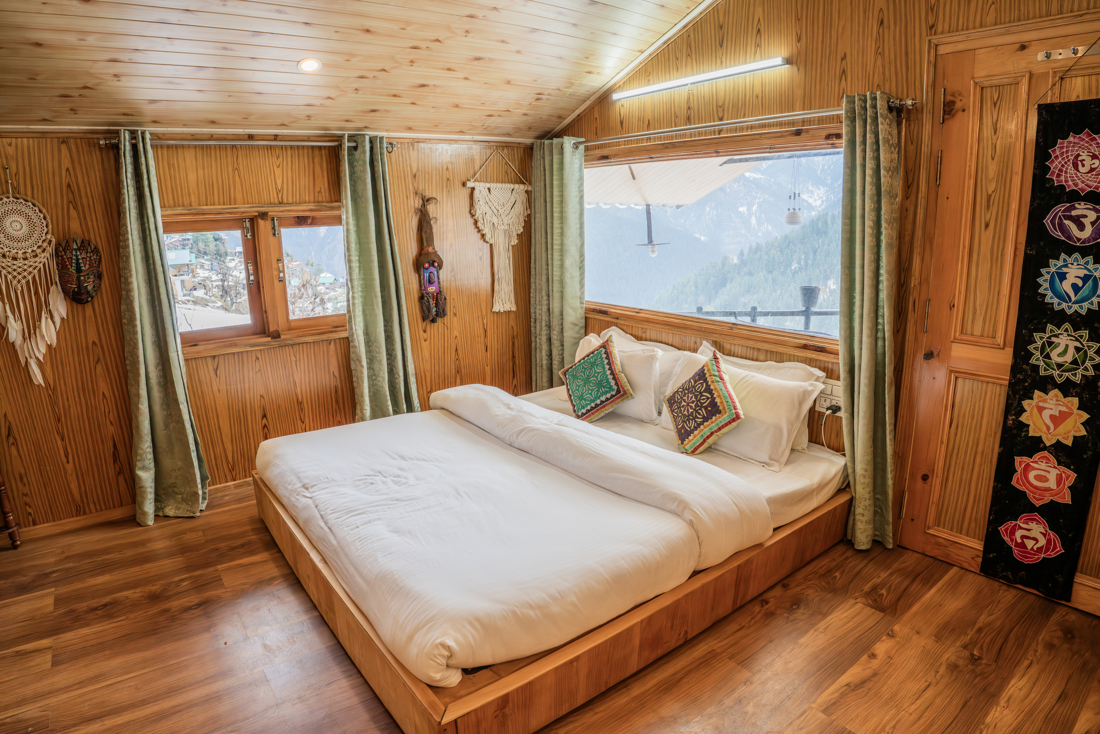 Bed made in white linen with Himachali cushions, dreamcatchers on the pine wall and mountains through the window