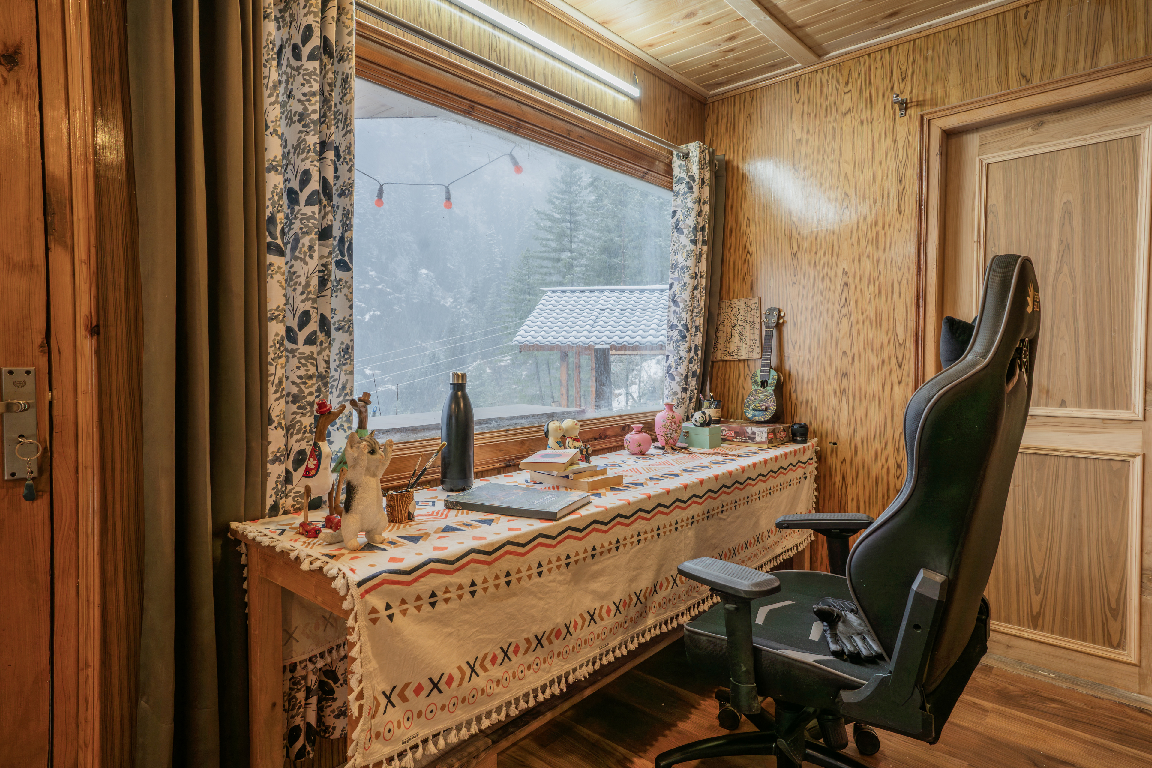 Study table set under a wide window with snow-covered deodars outside and a chair drawn up